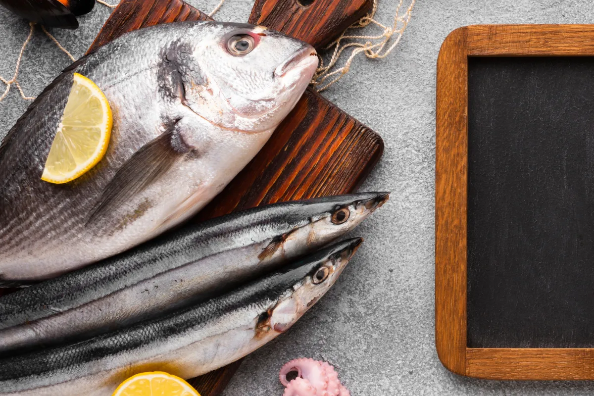 Vista superior de peixes frescos, tentáculos de polvo e fatias de limão em uma tábua de madeira ao lado de uma pequena lousa de menu, simbolizando o planejamento de um cardápio vendedor para frutos do mar.