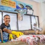 Funcionário sorridente usando luvas amarelas ao manipular peixes frescos em uma bancada de mercado, ilustrando diferentes modelos de negócio como peixaria e restaurante de frutos do mar.