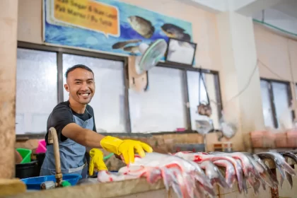 Funcionário sorridente usando luvas amarelas ao manipular peixes frescos em uma bancada de mercado, ilustrando diferentes modelos de negócio como peixaria e restaurante de frutos do mar.