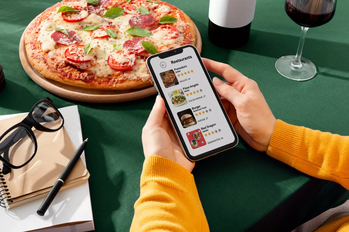Mãos segurando um smartphone exibindo um aplicativo de delivery com lista de restaurantes, com uma pizza ao fundo, ilustrando a importância da presença digital e em marketplaces para a divulgação da pizzaria