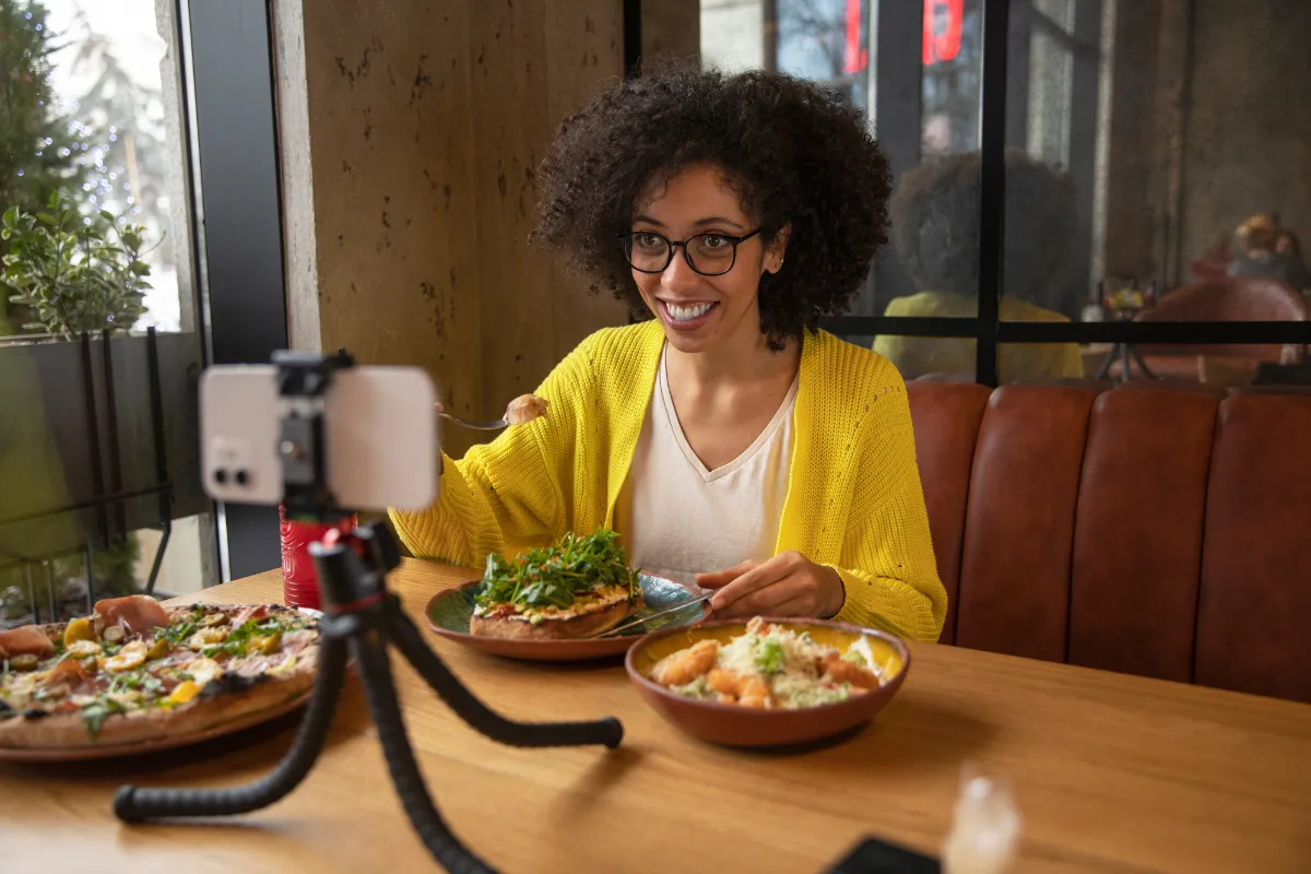 Mulher sorrindo sentada à mesa de um restaurante com pizzas e pratos, gravando um vídeo de si mesma com o celular em um tripé, conectando o marketing de influência à experiência de consumo no salão.