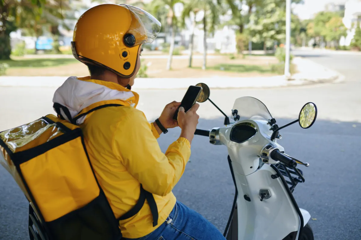 Motoboy visto de costas, vestindo jaqueta amarela e capacete da mesma cor, operando um smartphone enquanto está parado em sua moto. A imagem destaca o uso de tecnologia para gestão de pedidos, contextualizando o dilema entre contratar via aplicativos, MEI ou regime CLT.