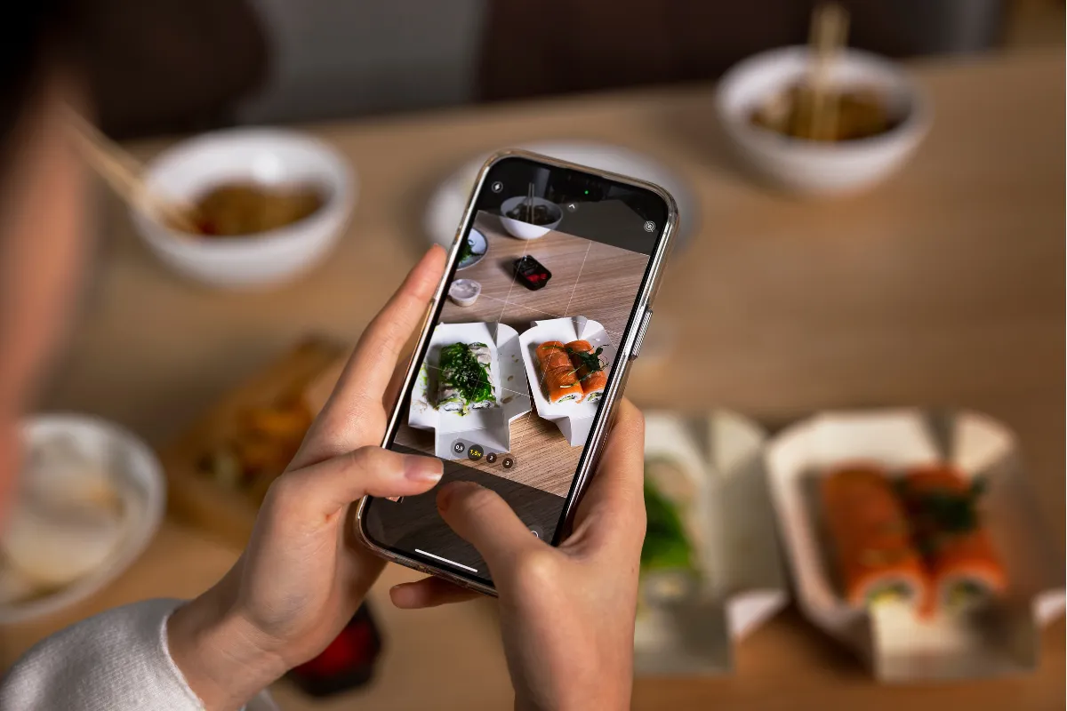 Mãos segurando um smartphone tirando foto de embalagens de delivery de sushi em uma mesa de madeira, ilustrando a produção de fotos atrativas ("food porn") para anúncios no Instagram e Meta Ads.
