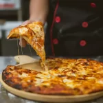 Close-up de uma mão levantando uma fatia de pizza com queijo derretido esticando, um exemplo clássico de imagem estilo 'food porn' ideal para propaganda de pizzaria nas redes sociais.