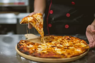 Close-up de uma mão levantando uma fatia de pizza com queijo derretido esticando, um exemplo clássico de imagem estilo 'food porn' ideal para propaganda de pizzaria nas redes sociais.