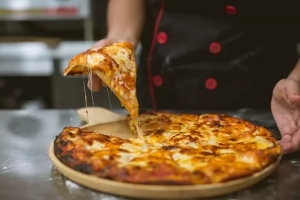 Close-up de uma mão levantando uma fatia de pizza com queijo derretido esticando, um exemplo clássico de imagem estilo 'food porn' ideal para propaganda de pizzaria nas redes sociais.