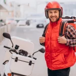 Entregador em pé ao lado de sua moto branca, sorrindo para a câmera. Ele veste um colete e capacete vermelhos e carrega uma mochila de entrega térmica nas costas. A imagem evoca a transparência de preços e a prontidão do serviço terceirizado para o mercado de 2026.