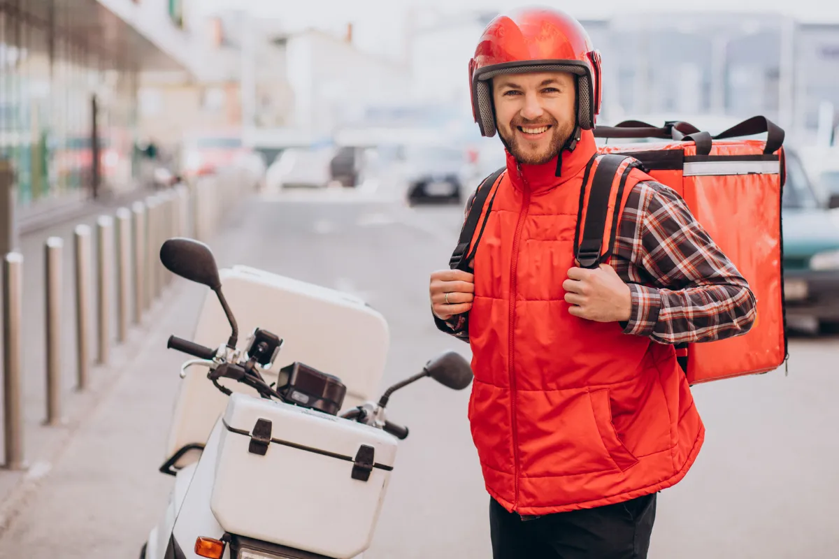 Entregador em pé ao lado de sua moto branca, sorrindo para a câmera. Ele veste um colete e capacete vermelhos e carrega uma mochila de entrega térmica nas costas. A imagem evoca a transparência de preços e a prontidão do serviço terceirizado para o mercado de 2026.