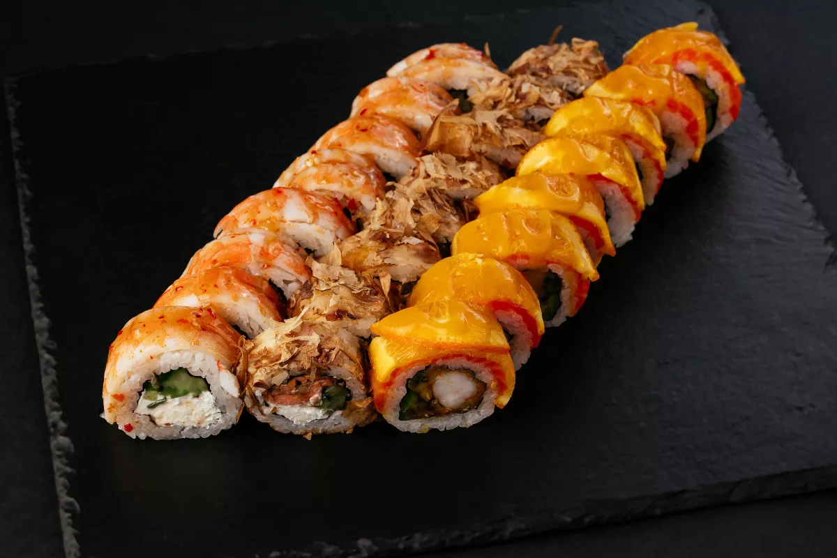 Sequência de Hot Roll (sushi frito) disposta sobre uma pedra preta, com recheio de salmão e cream cheese, cobertos com molho tarê e alguns finalizados com um toque de molho especial amarelo e cebolinha.