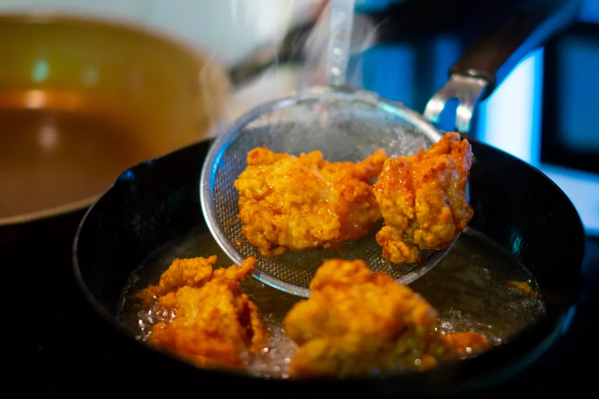 Pedaços de Karaage (frango frito japonês) sendo retirados de uma panela de óleo quente com uma escumadeira de metal, mostrando a textura dourada, crocante e suculenta do empanado típico.