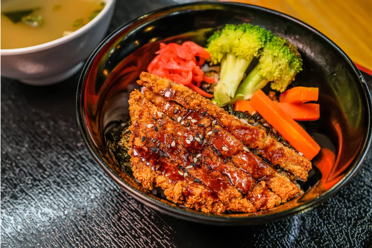 Tigela de Katsudon vista de cima, destacando o lombo de porco empanado e frito (tonkatsu) fatiado sobre uma cama de arroz e algas nori, finalizado com molho escuro e gergelim, acompanhado de brócolis, cenoura e gengibre em conserva.