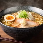 Tigela fumegante de Lámen japonês tradicional com caldo marrom, macarrão, fatias de carne de porco (chashu), meio ovo cozido com gema mole, broto de feijão e cebolinha picada sobre uma mesa de madeira.
