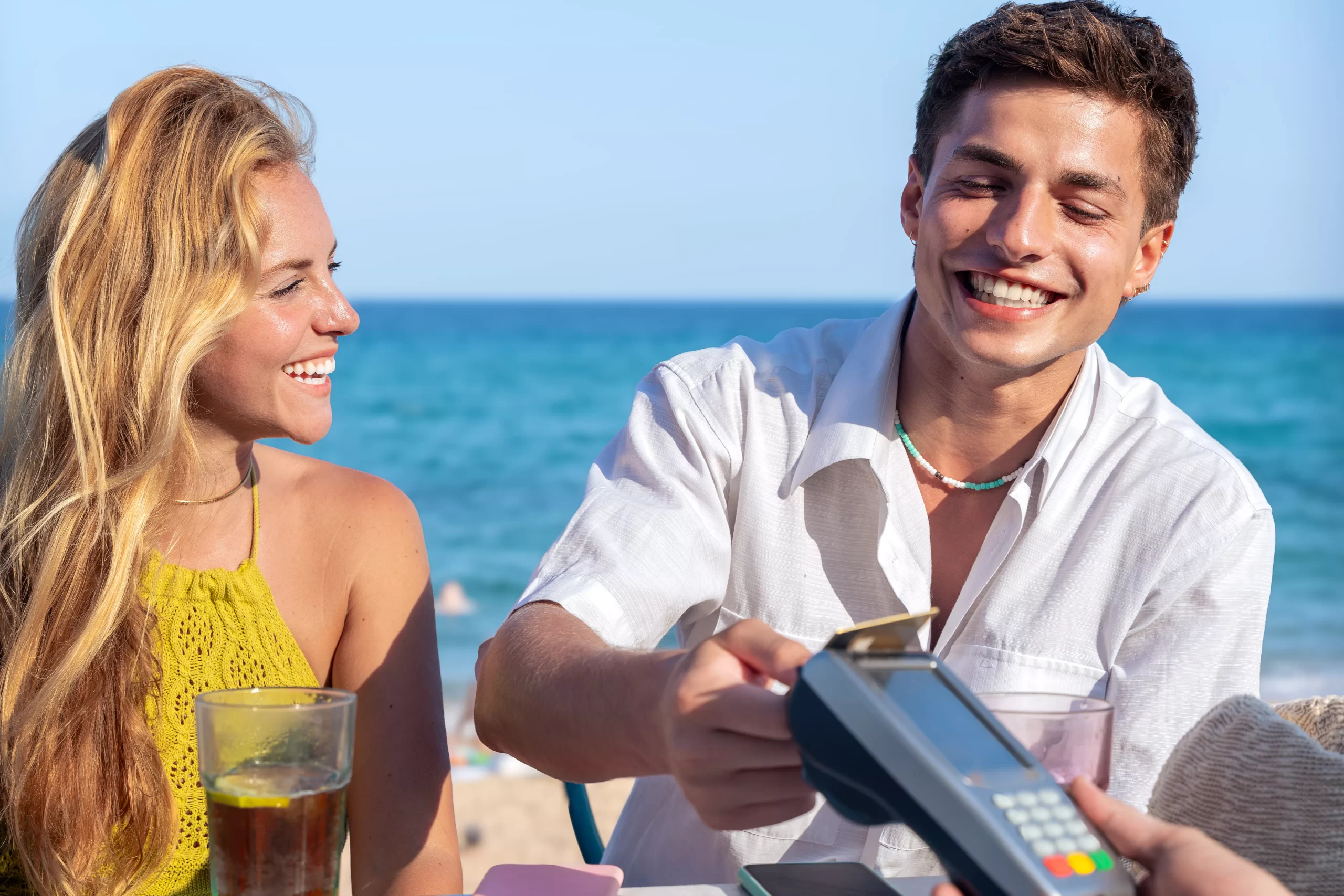 A imagem mostra um casal jovem sentado à beira-mar, em um ambiente típico de praia. Eles estão relaxados e sorridentes, sentados em cadeiras, com o oceano azul ao fundo, transmitindo um clima de verão, lazer e descontração. O rapaz, vestindo camisa branca leve, está realizando um pagamento com cartão em uma maquininha, segurando o cartão próximo ao equipamento. Ele aparenta estar confortável e feliz durante a ação. Ao lado dele, a mulher, com blusa amarela, observa a cena sorrindo. Ambos têm bebidas sobre a mesa, reforçando o contexto de consumo em um quiosque ou bar de praia.