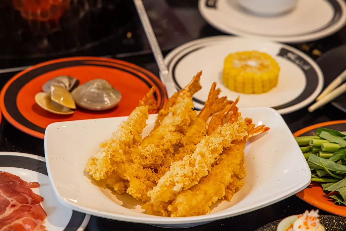 Porção de Tempurá de camarão servida em um prato branco, mostrando camarões grandes empanados em uma massa leve e extremamente crocante, com outros pratos orientais e acompanhamentos ao fundo.