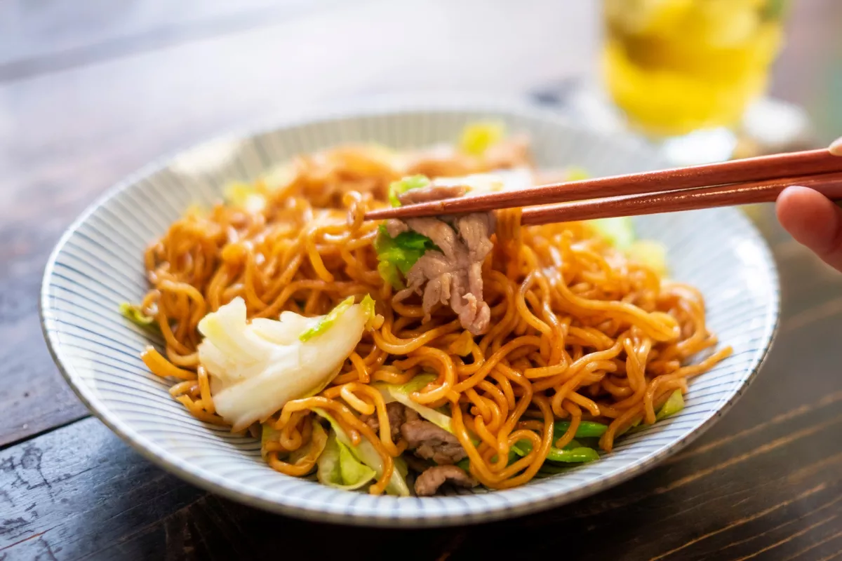 Prato de Yakissoba tradicional com macarrão frito refogado, pedaços de carne bovina, acelga e legumes ao molho shoyu, sendo degustado com o uso de hashis (palitinhos de madeira).