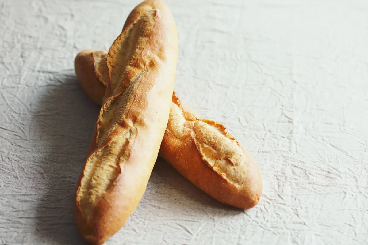 Duas baguetes francesas tradicionais cruzadas sobre um tecido de linho claro. Os pães são longos, com casca rústica, crocante e cortes diagonais dourados ao longo do comprimento.
