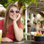 Mulher sorridente em um restaurante ao ar livre durante o verão de 2026, segurando um drink de frutas amarelas refrescante. À mesa, uma tigela de frutas tropicais frescas, ilustrando o conceito de luxo acessível e frescor no setor de food service.