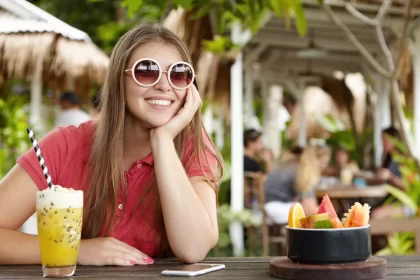 Mulher sorridente em um restaurante ao ar livre durante o verão de 2026, segurando um drink de frutas amarelas refrescante. À mesa, uma tigela de frutas tropicais frescas, ilustrando o conceito de luxo acessível e frescor no setor de food service.