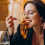 Mulher jovem de óculos sorrindo enquanto saboreia um prato de massa artesanal com molho de tomate em um restaurante, ilustrando a satisfação do cliente com um cardápio bem planejado.