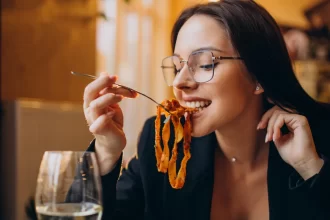 Mulher jovem de óculos sorrindo enquanto saboreia um prato de massa artesanal com molho de tomate em um restaurante, ilustrando a satisfação do cliente com um cardápio bem planejado.