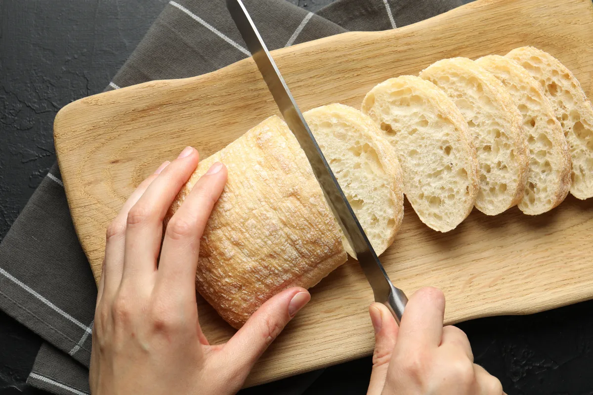 Mãos cortando uma ciabatta italiana em fatias sobre uma tábua de madeira. O pão tem formato achatado e o miolo revela os grandes alvéolos (buracos) característicos da alta hidratação da massa