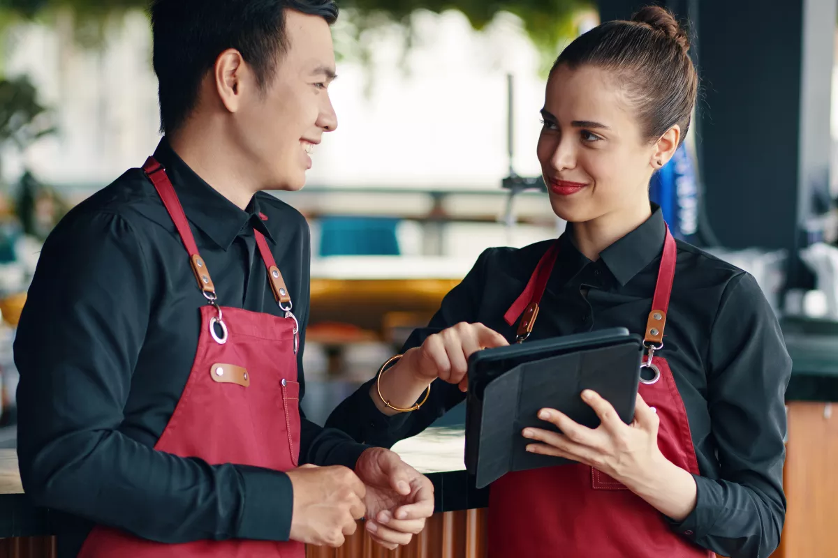 Dois atendentes de bar sorridentes, usando uniformes pretos e aventais vermelhos, operam uma comanda eletrônica em um tablet. A ferramenta substitui o papel, garantindo agilidade no atendimento e eliminando erros de pedido no envio direto para a produção.