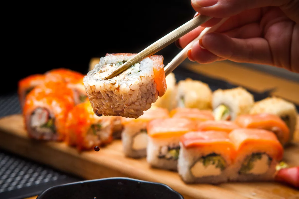Mão segurando um sushi com hashi sobre uma barca de combinados japoneses, ilustrando o rodízio de comida japonesa.
