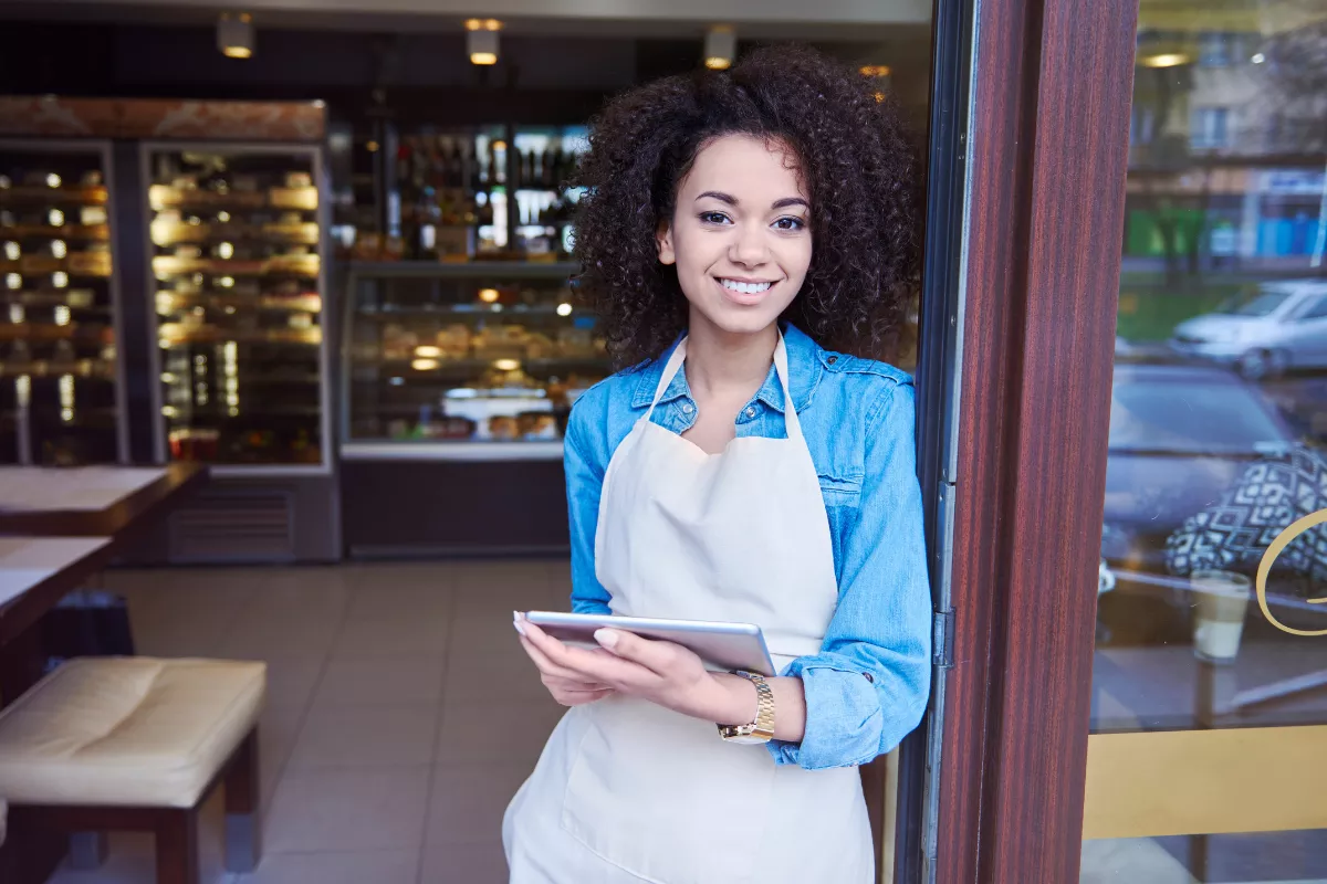 Jovem dona de restaurante sorridente, usando avental branco sobre camisa jeans, segurando um tablet na entrada da sua cafeteria. A imagem ilustra o uso da tecnologia para receber pedidos e organizar a operação, refletindo a facilidade de ter a gestão do delivery integrada na palma da mão.