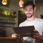 Gestor de restaurante sorrindo ao consultar dados no tablet, representando a facilidade de fazer análises preditivas com o sistema ControleNaMão em um ambiente de bar descontraído.