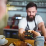 Close das mãos de um cliente realizando pagamento por aproximação em máquina de cartão no balcão, com o caixa ao fundo, agilizando o processo de Frente de Caixa e PDV.
