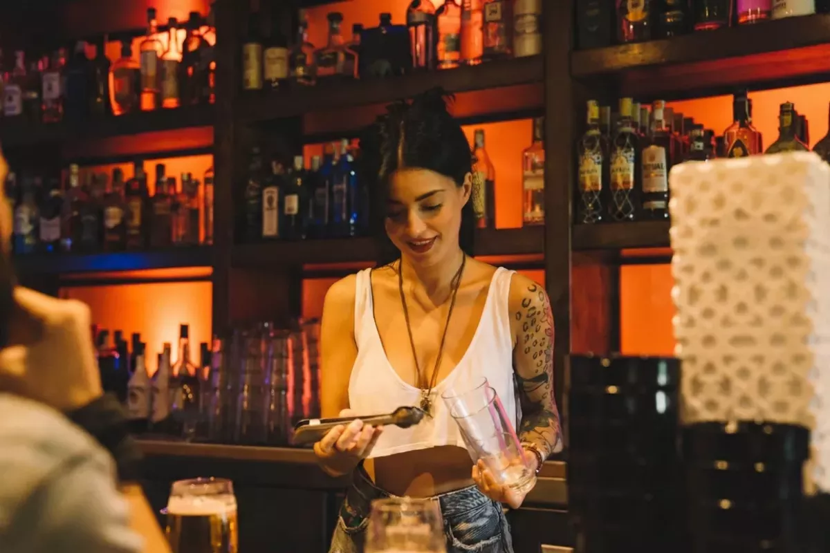 Bartender feminina preparando um drinque em um bar com prateleiras repletas de garrafas ao fundo sob luz âmbar. A cena ilustra a importância da gestão de estoque e ficha técnica para o controle rigoroso de insumos e cálculo de CMV.