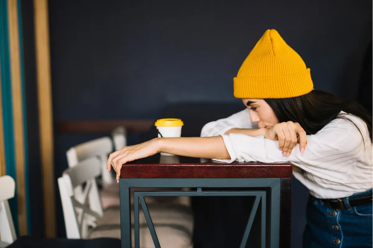 Mulher jovem com gorro amarelo e expressão de cansaço e desânimo, com a cabeça apoiada nos braços sobre uma mesa alta em um ambiente de cafeteria, ilustrando o esgotamento mental.