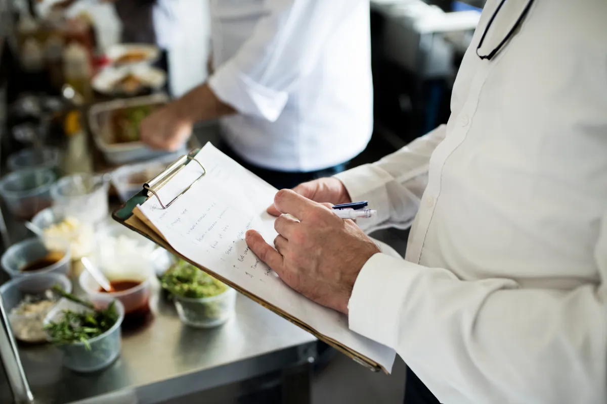 Close nas mãos de um chef de cozinha escrevendo em uma prancheta, com ingredientes organizados em potes e a equipe trabalhando ao fundo em uma cozinha profissional movimentada.
