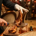 Garçom com luva branca cortando uma costela assada em uma tábua, no contexto de um rodízio de carnes.