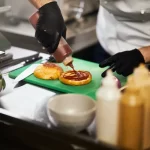Foto em primeiro plano das mãos de um cozinheiro usando luvas pretas enquanto finaliza a montagem de um hambúrguer em uma cozinha profissional. Ele aplica molho ketchup em um pão selado sobre uma tábua verde, ilustrando a padronização e agilidade característica do modelo de fast food.