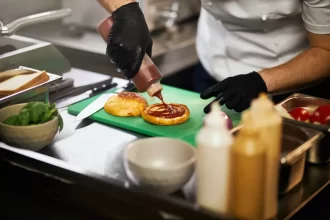 Foto em primeiro plano das mãos de um cozinheiro usando luvas pretas enquanto finaliza a montagem de um hambúrguer em uma cozinha profissional. Ele aplica molho ketchup em um pão selado sobre uma tábua verde, ilustrando a padronização e agilidade característica do modelo de fast food.
