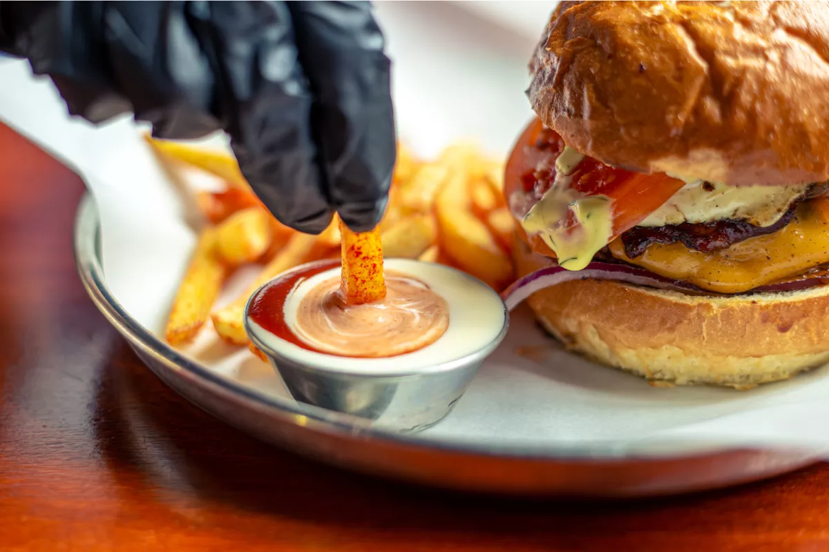 Imagem aproximada de um hambúrguer artesanal suculento com queijo derretido ao lado de batatas fritas crocantes. Uma mão com luva preta mergulha uma batata em um pequeno pote de molho rosé e ketchup, representando o conceito de junk food focado em alta densidade calórica e palatabilidade.