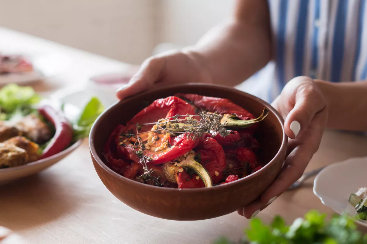 Mãos femininas segurando uma tigela de cerâmica rústica repleta de pimentões vermelhos assados e temperados com ervas frescas. Ao fundo, uma mesa posta com alimentos naturais, remetendo à filosofia do slow food que valoriza ingredientes orgânicos, produção local e o prazer de comer com calma.