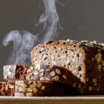 Pão 100% integral fatiado saindo fumaça, indicando frescor. O miolo é escuro, rico em fibras e sementes, com a crosta coberta por flocos de aveia, posicionado em um ambiente de luz suave.