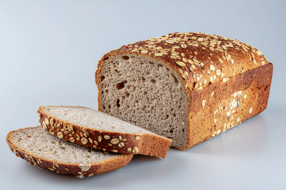 Pão de forma multigrãos fatiado, mostrando um miolo denso e nutritivo. A crosta superior é generosamente coberta por flocos de aveia e sementes diversas, reforçando o apelo saudável