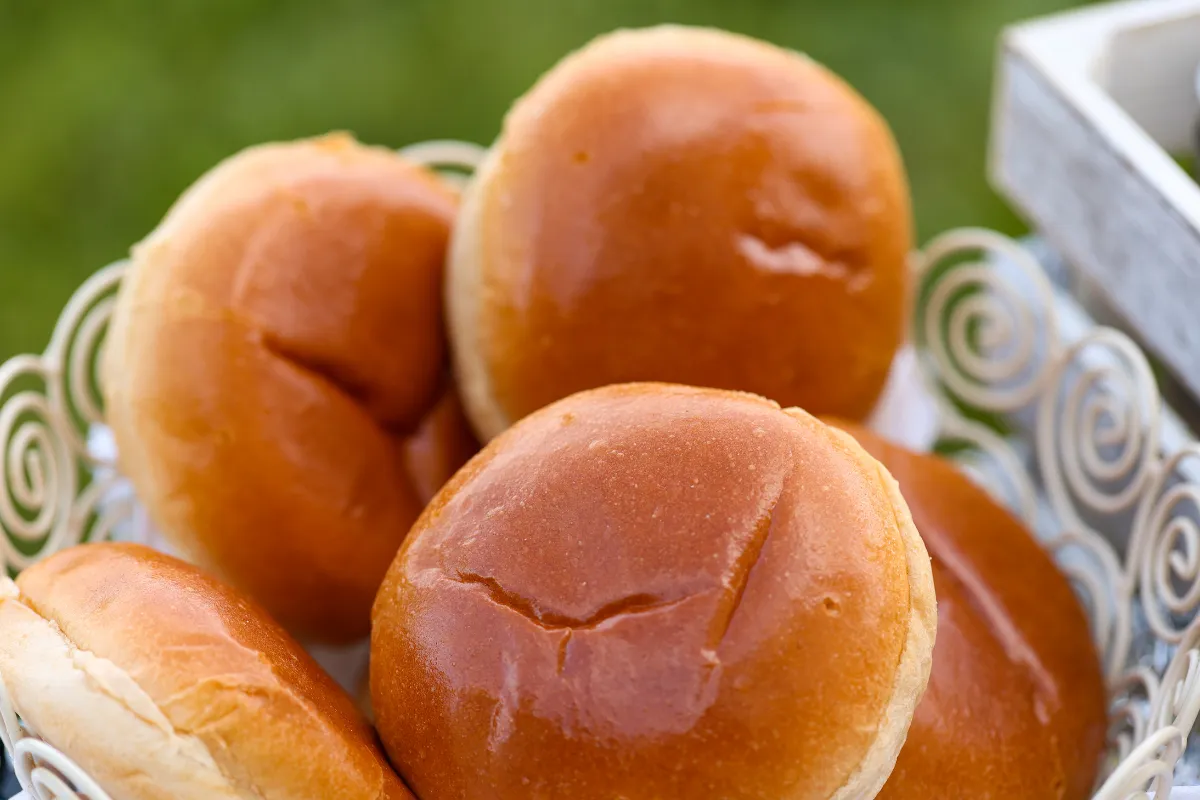 Cesta de metal branca com detalhes em espiral contendo vários pães de hambúrguer tipo brioche. Os pães possuem uma superfície dourada, lisa e brilhante, ideal para lanches gourmet