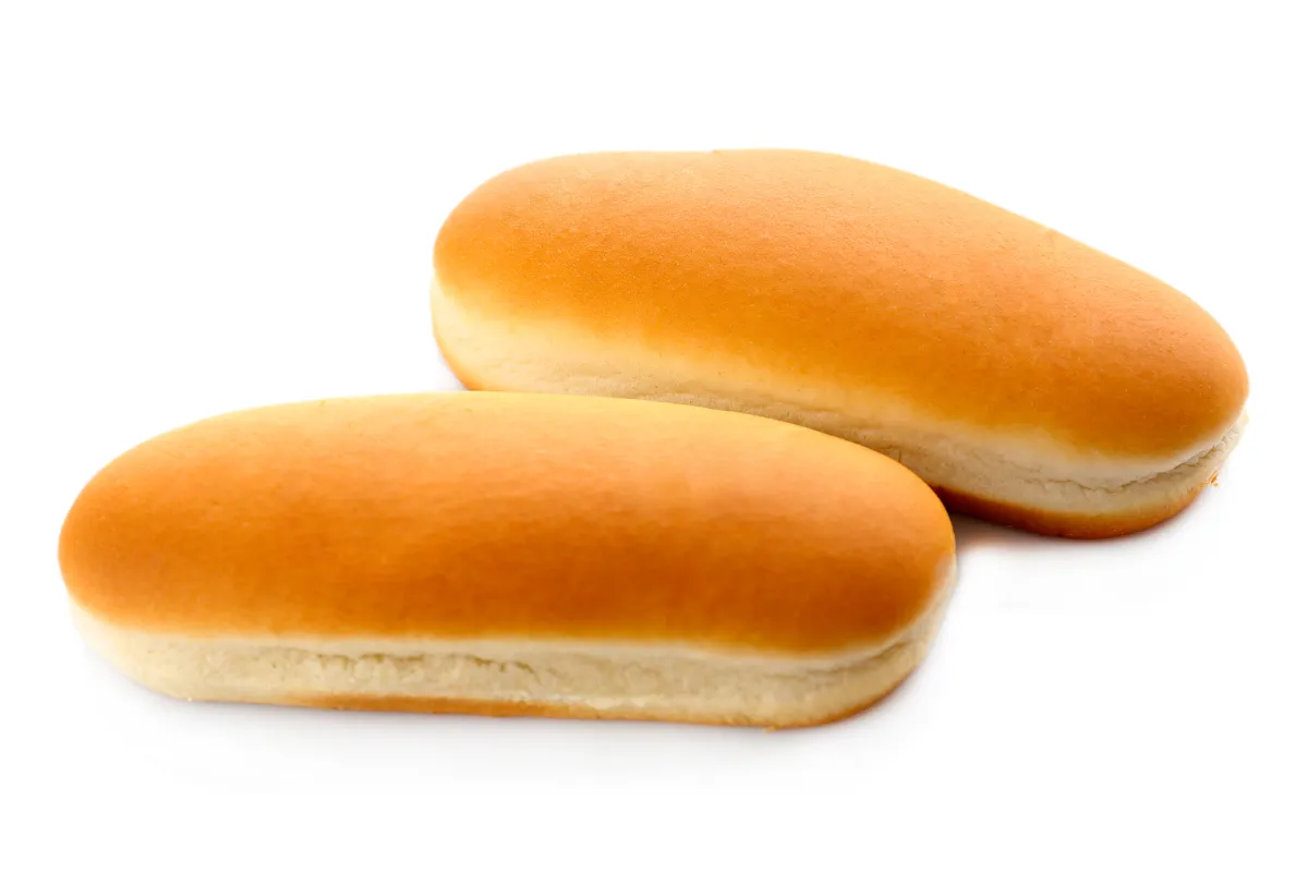 Dois pães de cachorro-quente alongados e macios dispostos sobre um fundo branco. Os pães apresentam uma coloração dourada uniforme e textura leve, típicos para montagem de lanches rápidos