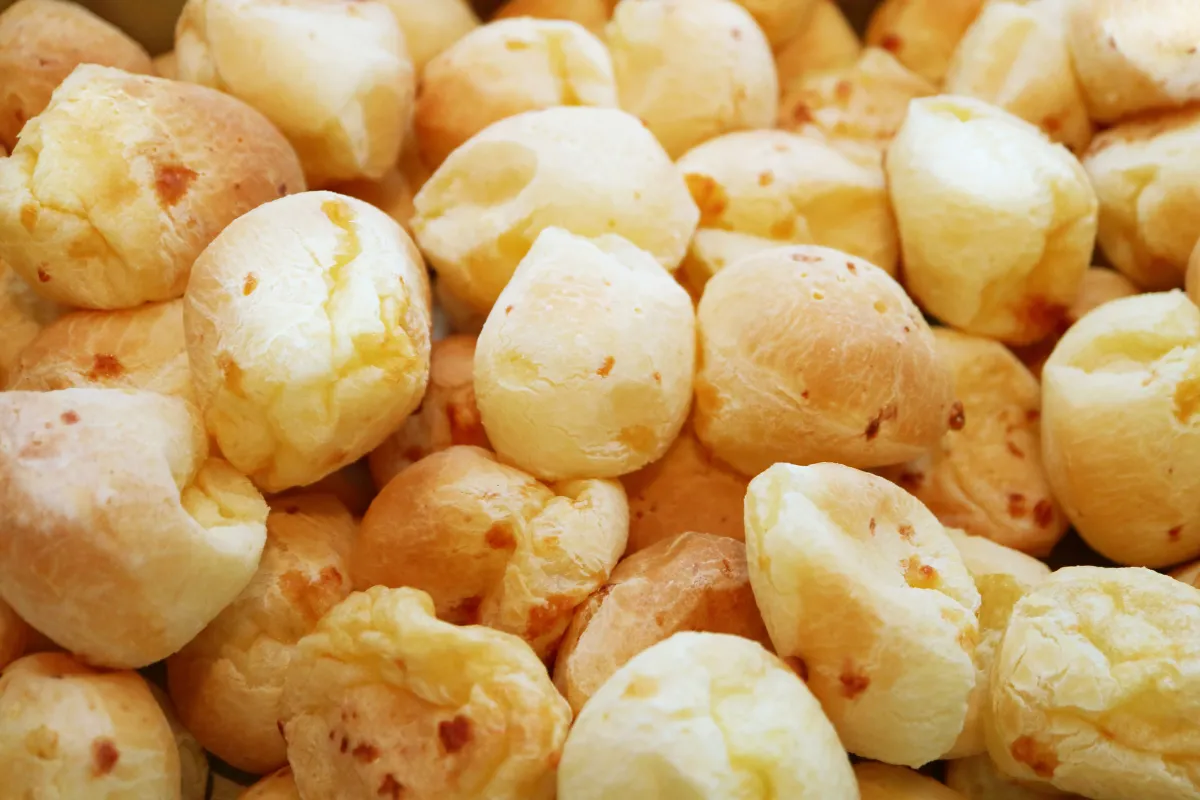 Grande quantidade de pães de queijo mineiros tradicionais, com formato arredondado, casca dourada e irregular, apresentando a textura típica de polvilho e queijo assado