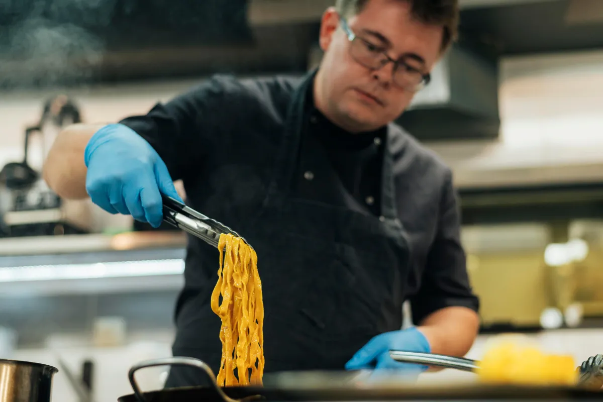 Chef de cozinha profissional usando luvas azuis e avental, utilizando um pegador para servir massa fresca longa (tagliatelle) saindo de uma panela com vapor, representando a operação e equipamentos de uma cozinha industrial.
