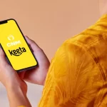 Mão segurando um smartphone que exibe na tela amarela os logotipos do ControleNaMão (CNM) e do aplicativo de delivery Keeta, simbolizando a integração tecnológica entre as plataformas.