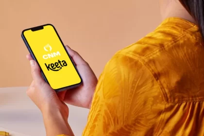 Mão segurando um smartphone que exibe na tela amarela os logotipos do ControleNaMão (CNM) e do aplicativo de delivery Keeta, simbolizando a integração tecnológica entre as plataformas.