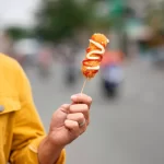 Homem vestindo jaqueta amarela segura um espetinho de aperitivo frito em frente a uma rua movimentada e desfocada. O petisco está decorado com ziguezagues de maionese e ketchup, ilustrando a praticidade de alimentação rápida durante os blocos de rua