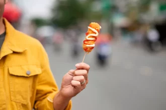 Homem vestindo jaqueta amarela segura um espetinho de aperitivo frito em frente a uma rua movimentada e desfocada. O petisco está decorado com ziguezagues de maionese e ketchup, ilustrando a praticidade de alimentação rápida durante os blocos de rua