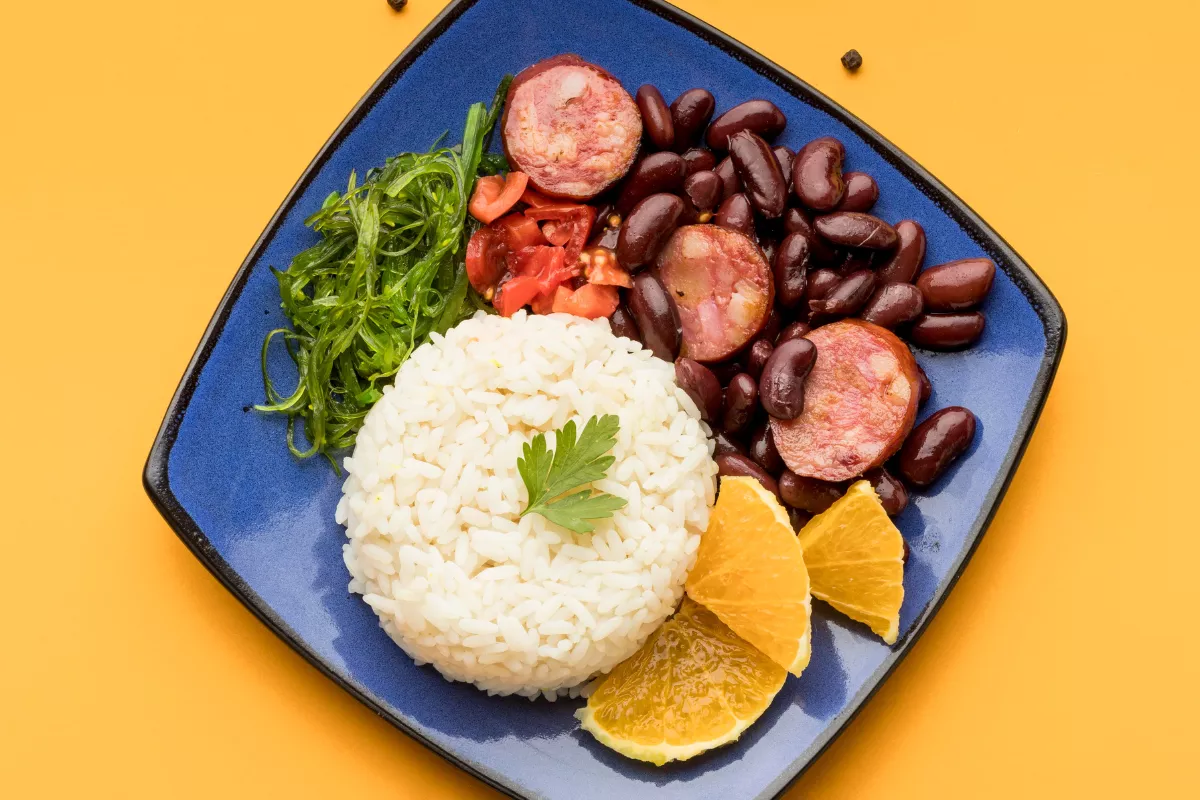 Vista superior de um prato azul contendo uma porção de feijoada brasileira com arroz branco, feijão preto com linguiça, couve refogada e fatias de laranja, posicionados sobre um fundo amarelo vibrante para destacar pratos reconfortantes pós-festa.