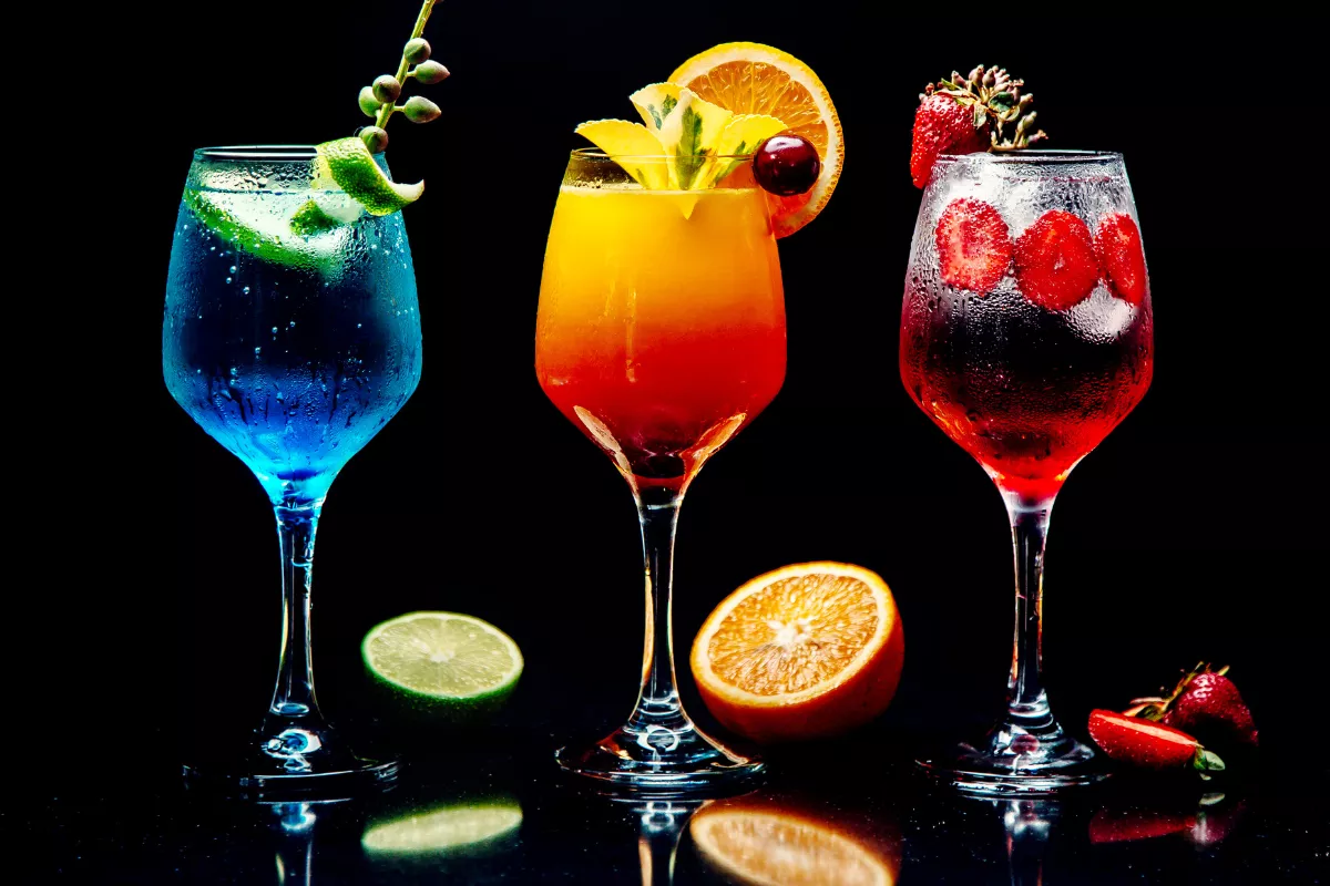 Três mocktails coloridos e decorados com frutas em taças de vidro sobre fundo escuro. Drinks sem álcool refrescantes (azul, laranja e vermelho) que são tendência de alta margem de lucro para o verão 2026.